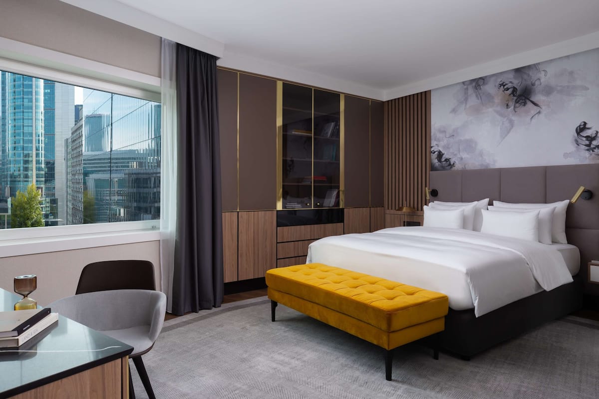 Habitación de hotel moderna con una cama grande, un reposapiés amarillo, un escritorio con una silla, una estantería y vistas a la ciudad.