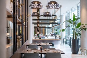 Un restaurant moderne doté d’une longue table à manger, de nombreuses chaises, de luminaires suspendus et d’une grande plante en pot.