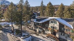 Un edificio de varios pisos con tejado cubierto de nieve, rodeado de altos pinos y una montaña al fondo.