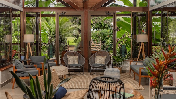 A spacious indoor area with large windows, wicker furniture, and potted plants. - Surf Synergy All-Inclusive Surf Retreat (Jacó)