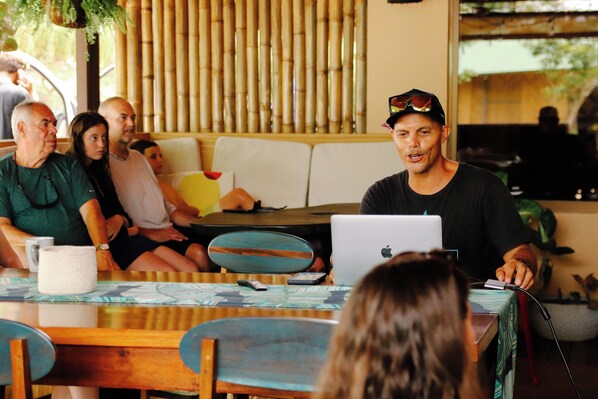 A group of people seated around a table with a laptop, in a room with bamboo blinds and a sofa.