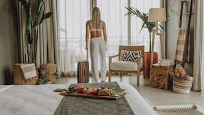 A hotel room with a bed, a dining table with food, a suitcase, a chair, a lamp, and a large window with curtains. - Surf Synergy All-Inclusive Surf Retreat (Jacó)