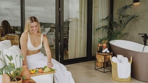 A woman seated on a white sofa in a room with a large bathtub, a wooden side table with a towel, and potted plants. - Surf Synergy All-Inclusive Surf Retreat (Jacó)