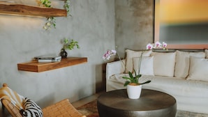 A modern living room with a sofa, a round coffee table, a woven chair, and a wooden shelf with plants. - Surf Synergy All-Inclusive Surf Retreat (Jacó)