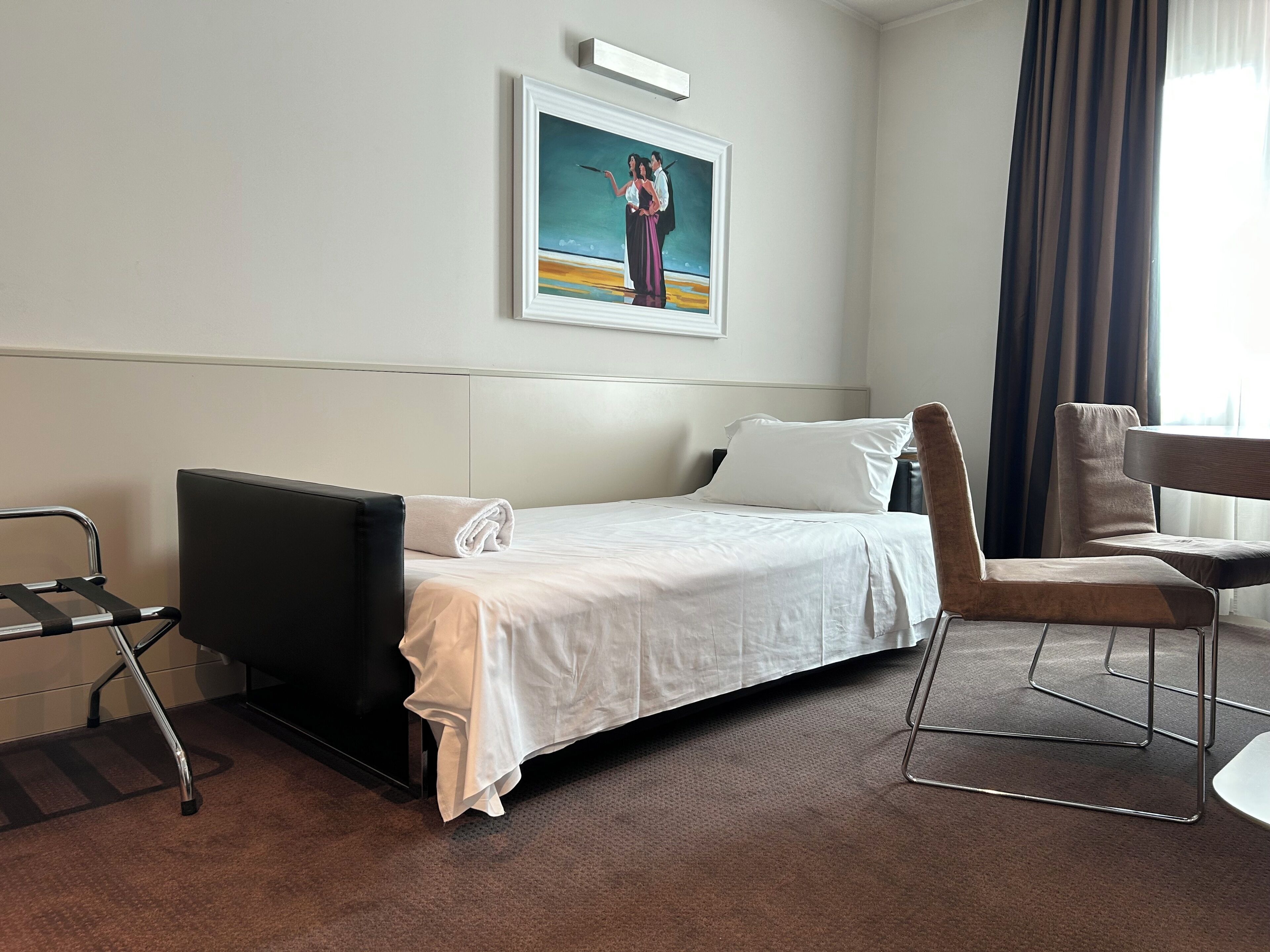 Ett hotellrum med en säng, en stol, ett skrivbord, en tavla på väggen och ett fönster med gardiner.