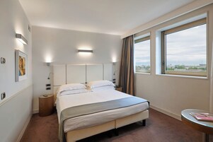 Habitación de hotel con una cama grande, una mesita de noche pequeña, una ventana con cortinas y vistas a la ciudad.