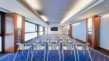 A conference room with rows of white chairs, a blue carpet with a pattern, and a sign that reads "PLAZA VENICE MESTRE."