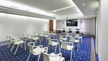 Un salón de conferencias con filas de sillas blancas, una alfombra azul con estampado floral y una pantalla grande que muestra