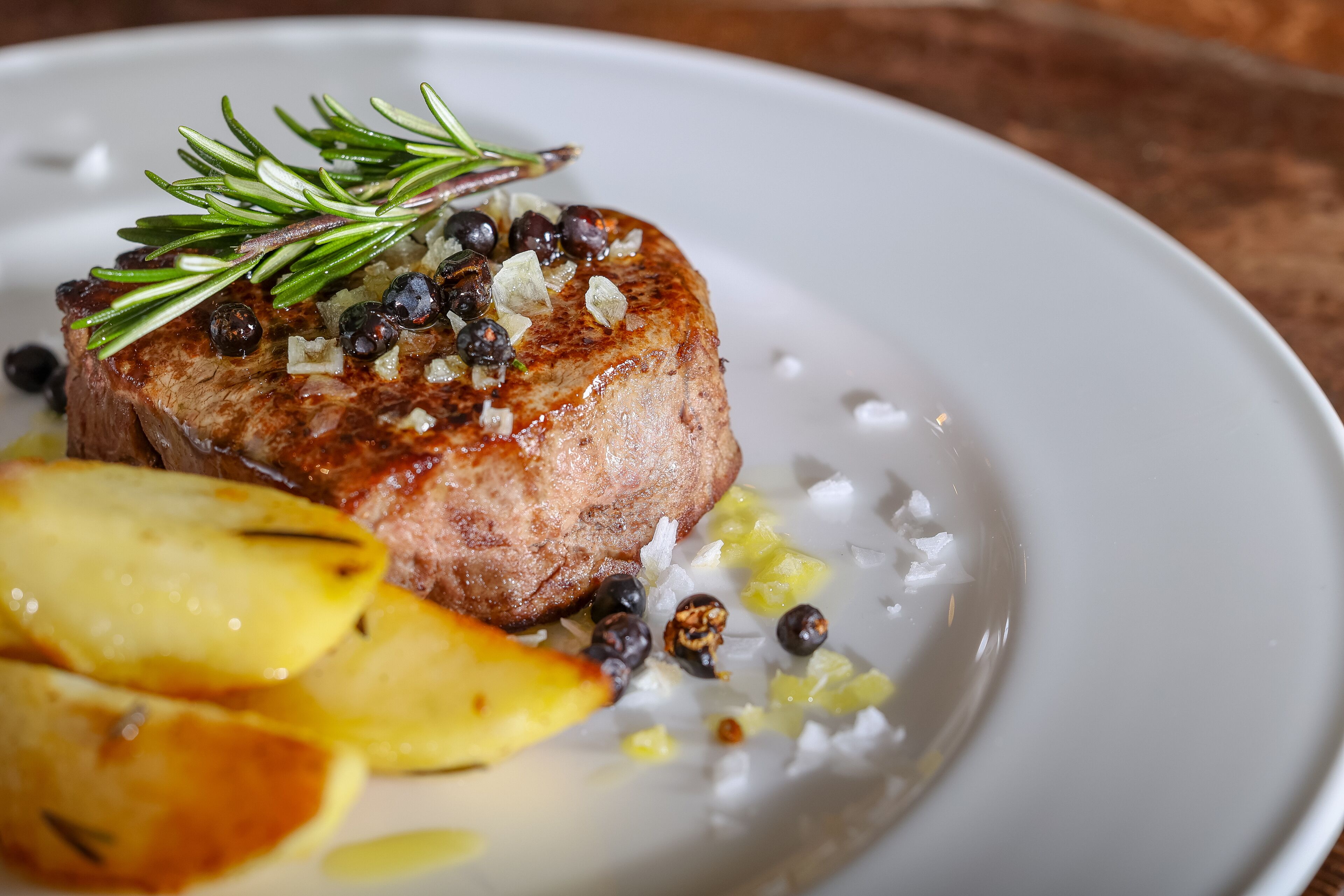 A plate with a grilled steak, roasted potatoes, and a garnish of rosemary and black peppercorns.