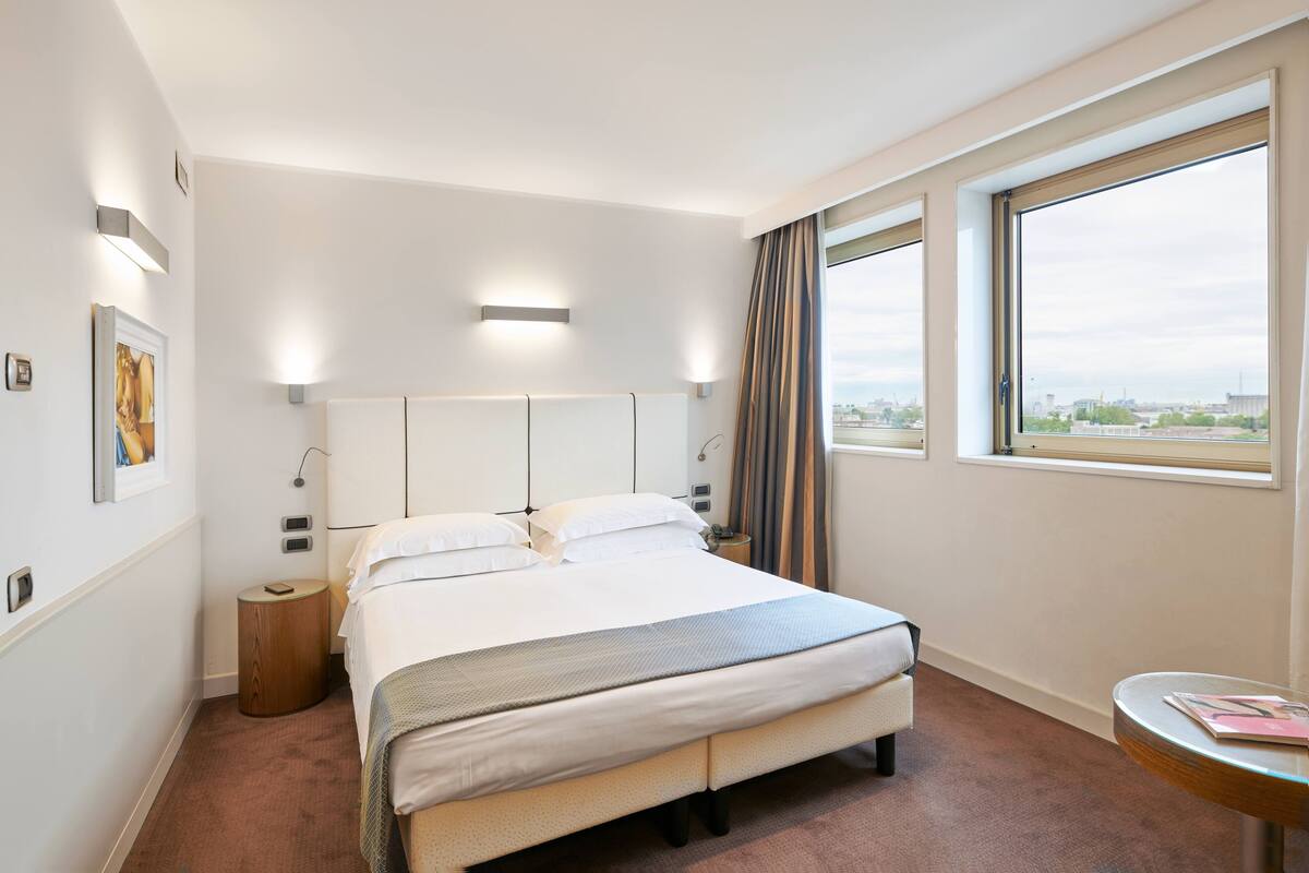 Une chambre d’hôtel avec un grand lit, une table de chevet, une petite table d’appoint ronde en bois, une fenêtre avec des rideaux et une vue sur la ville.