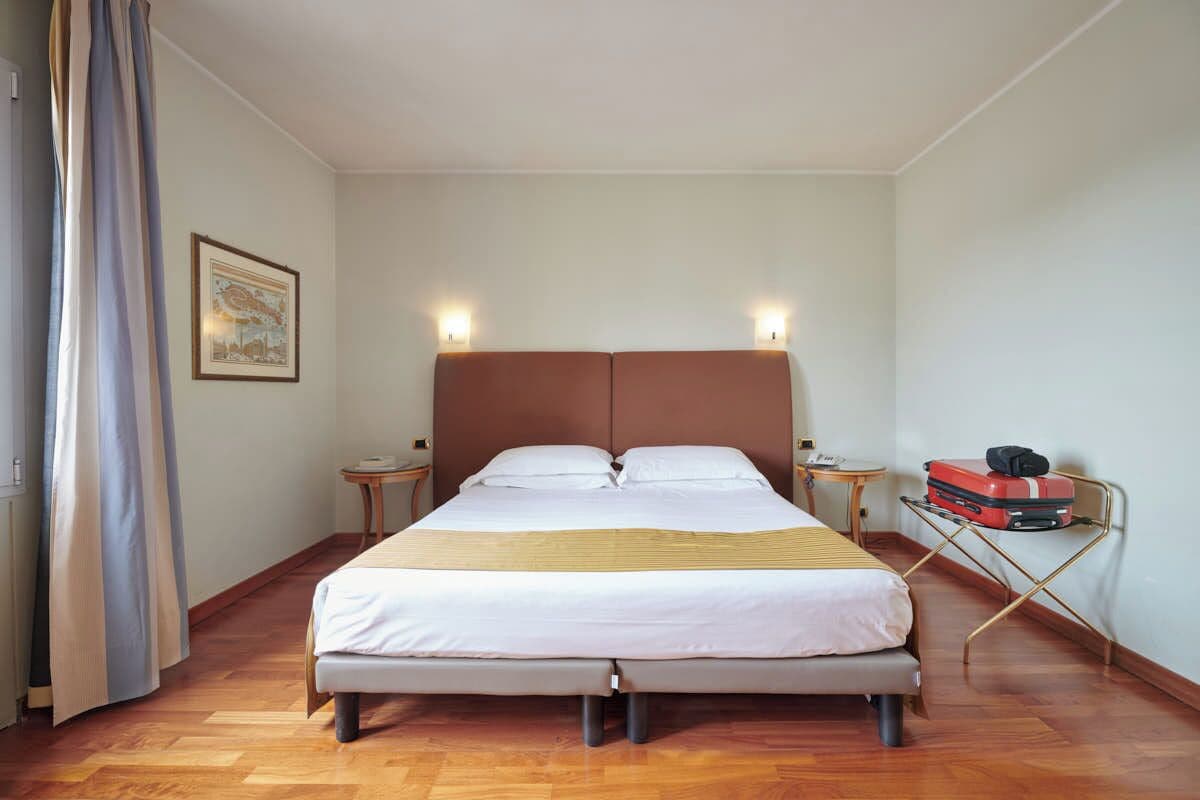 Uma cama bem arrumada com cabeceira, duas mesas de cabeceira, uma luminária na parede e uma mala sobre um suporte.