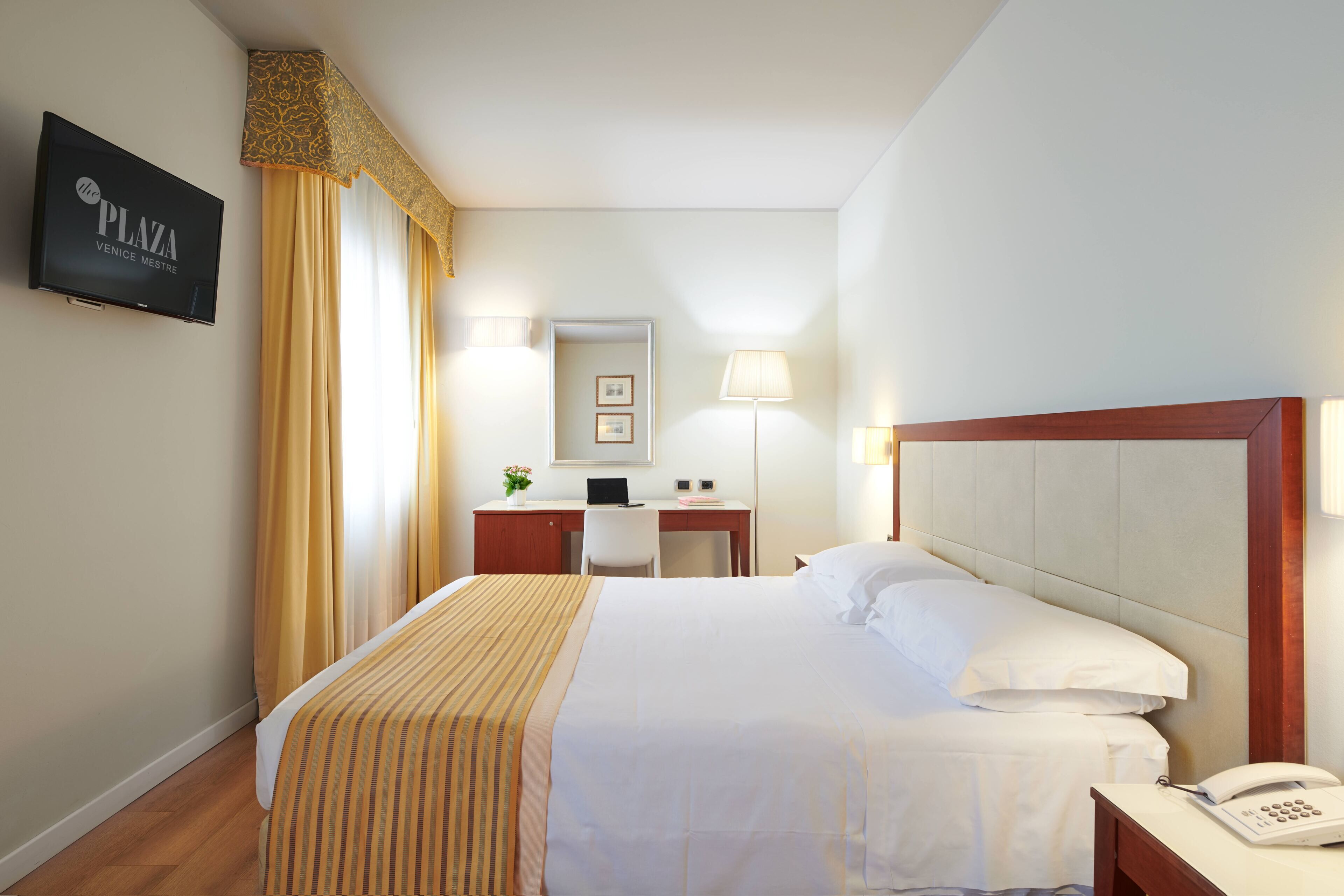Ein Hotelzimmer mit Bett, Schreibtisch, Stuhl, einem an der Wand befestigten Fernseher und einem Telefon auf dem Nachttisch.