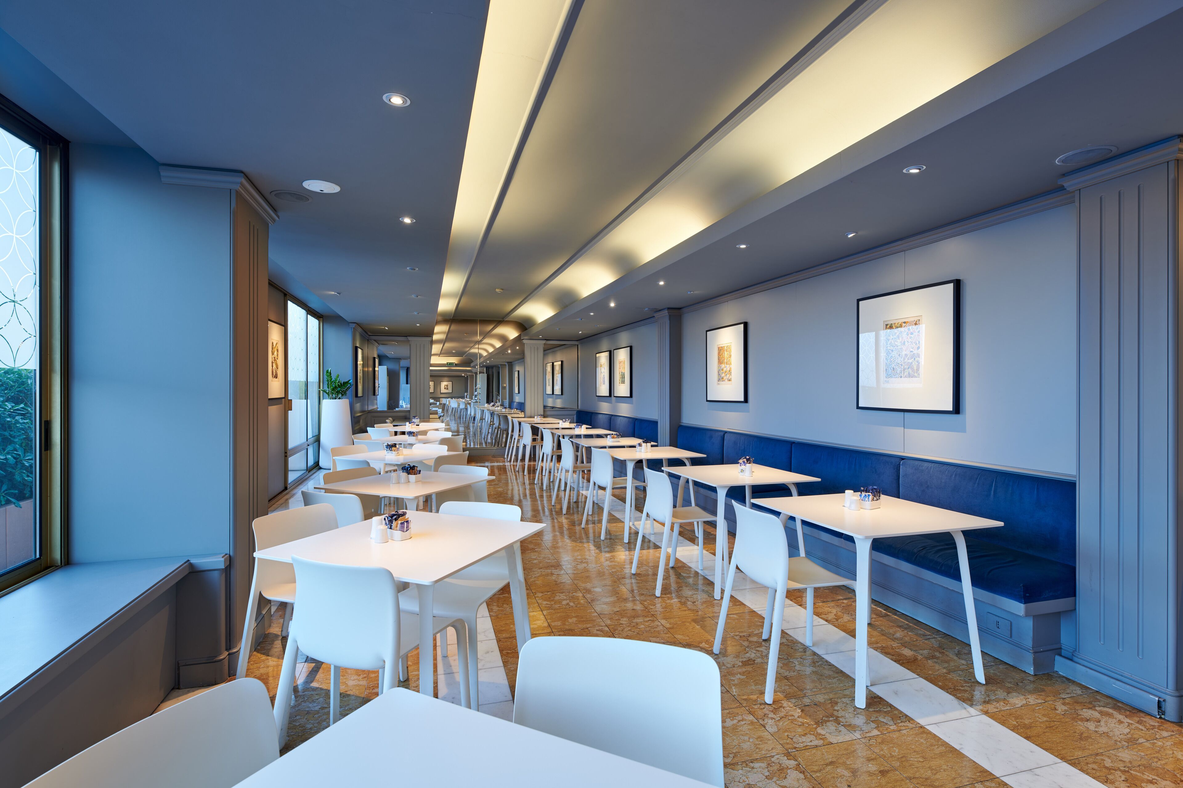 Un espace de restauration moderne avec des tables et des chaises blanches, des œuvres d’art encadrées accrochées aux murs et de grandes fenêtres sur un des côtés.