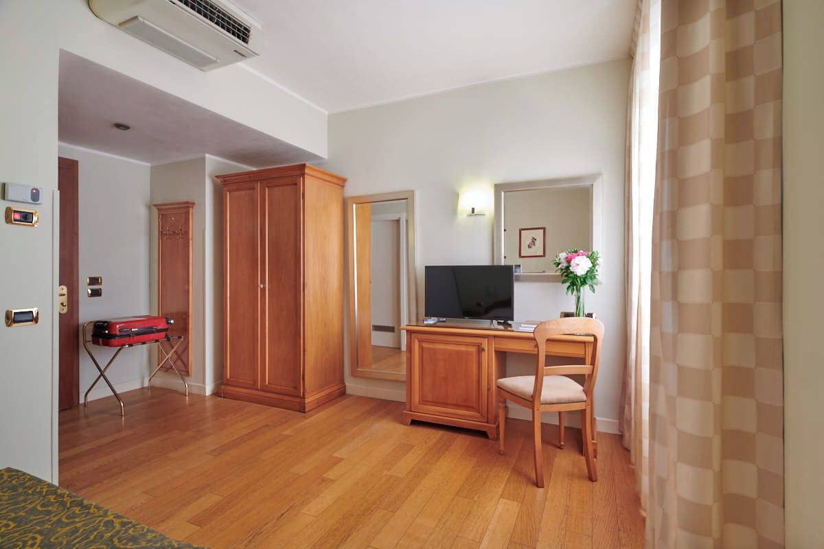 Quarto com piso de madeira, uma escrivaninha com computador, uma cadeira, um guarda-roupa e uma mala vermelha.