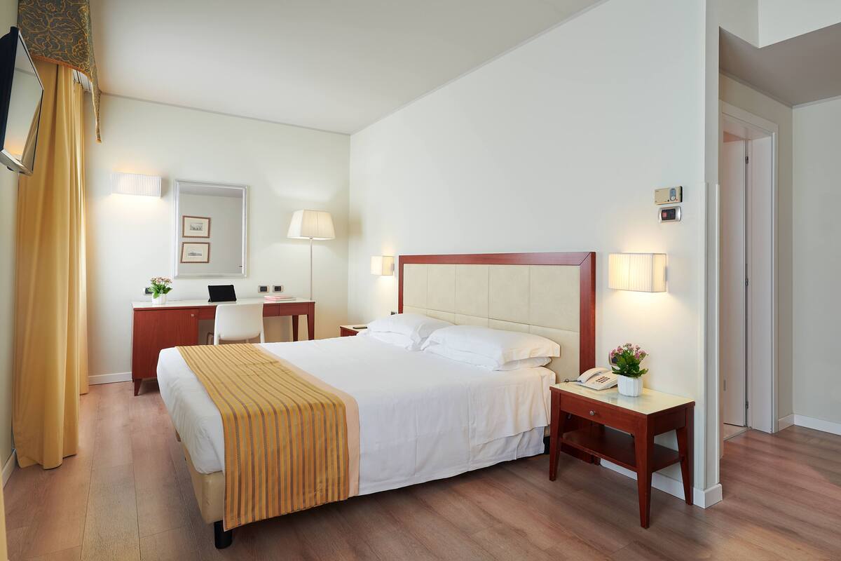 Une chambre d’hôtel avec un grand lit, des tables de chevet, un bureau et une chaise. Il y a un miroir au mur, une lampe et un téléphone sur le bureau.