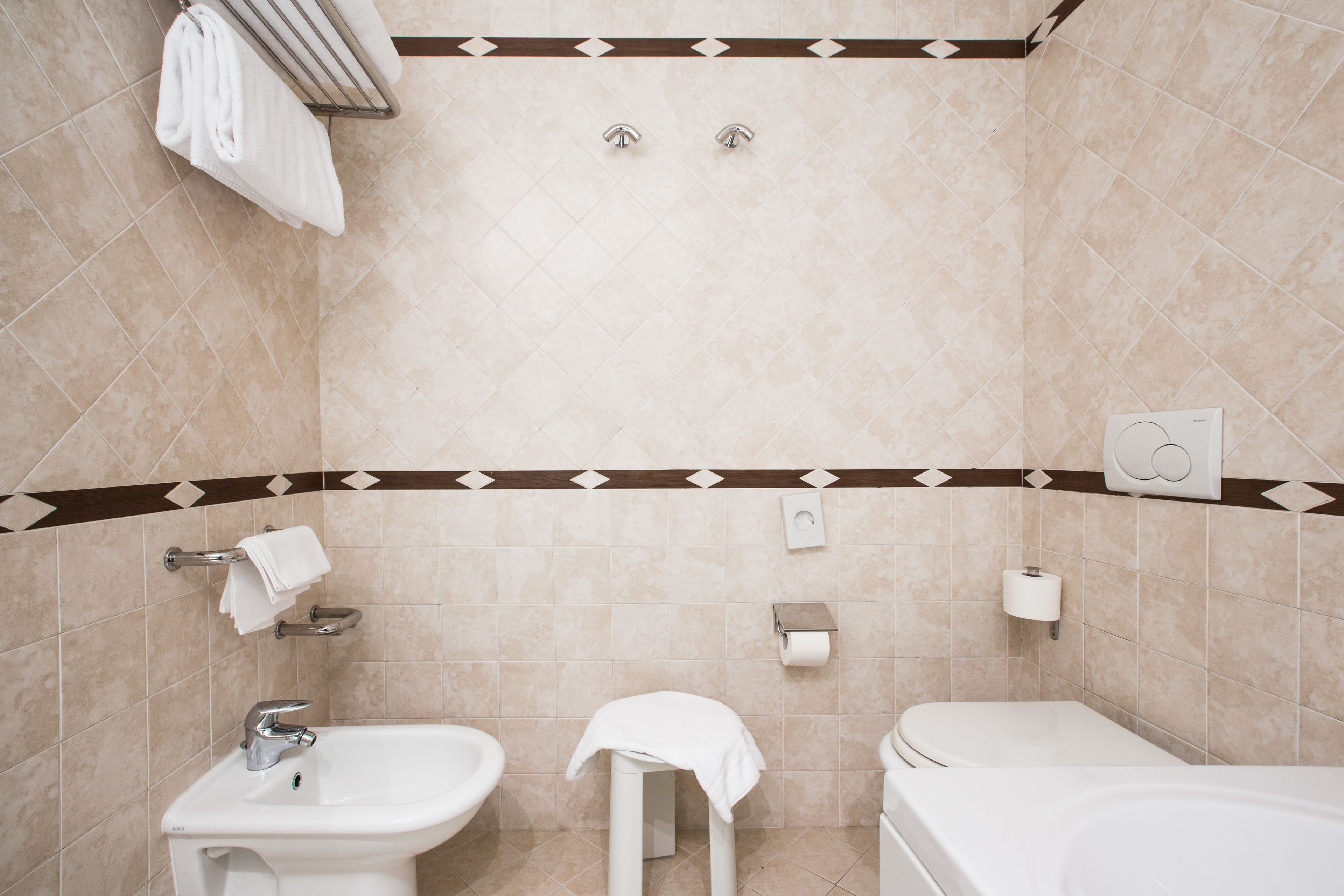 Un baño con lavabo, inodoro y bañera. Hay toallas colgadas en un perchero, y se ve un dispensador de papel higiénico en la pared.