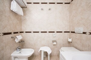 Un baño con lavabo, inodoro y bañera. Hay toallas colgadas en un perchero, y se ve un dispensador de papel higiénico en la pared.
