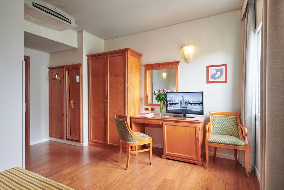 Quarto de hotel com secretária de madeira, duas cadeiras, televisão, espelho e guarda-roupa.