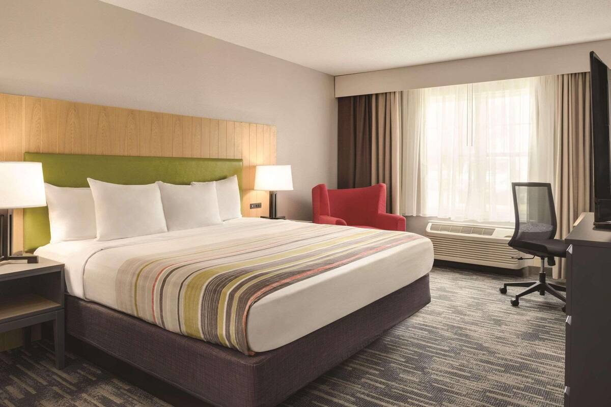 A hotel room with a large bed, a red chair, a desk with a chair, and a large window with curtains.