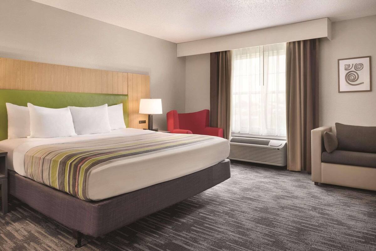 A hotel room with a large bed, a red chair, a grey sofa, a window with curtains, and a wall-mounted artwork.