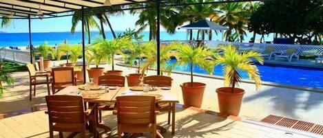 Un área de comedor al aire libre cubierta con vista a la piscina, mesas, sillas y plantas en macetas.