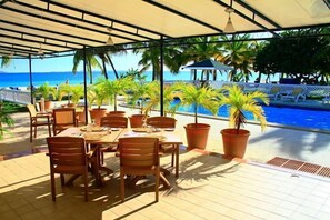 Un área de comedor al aire libre cubierta con vista a la piscina, mesas, sillas y plantas en macetas.