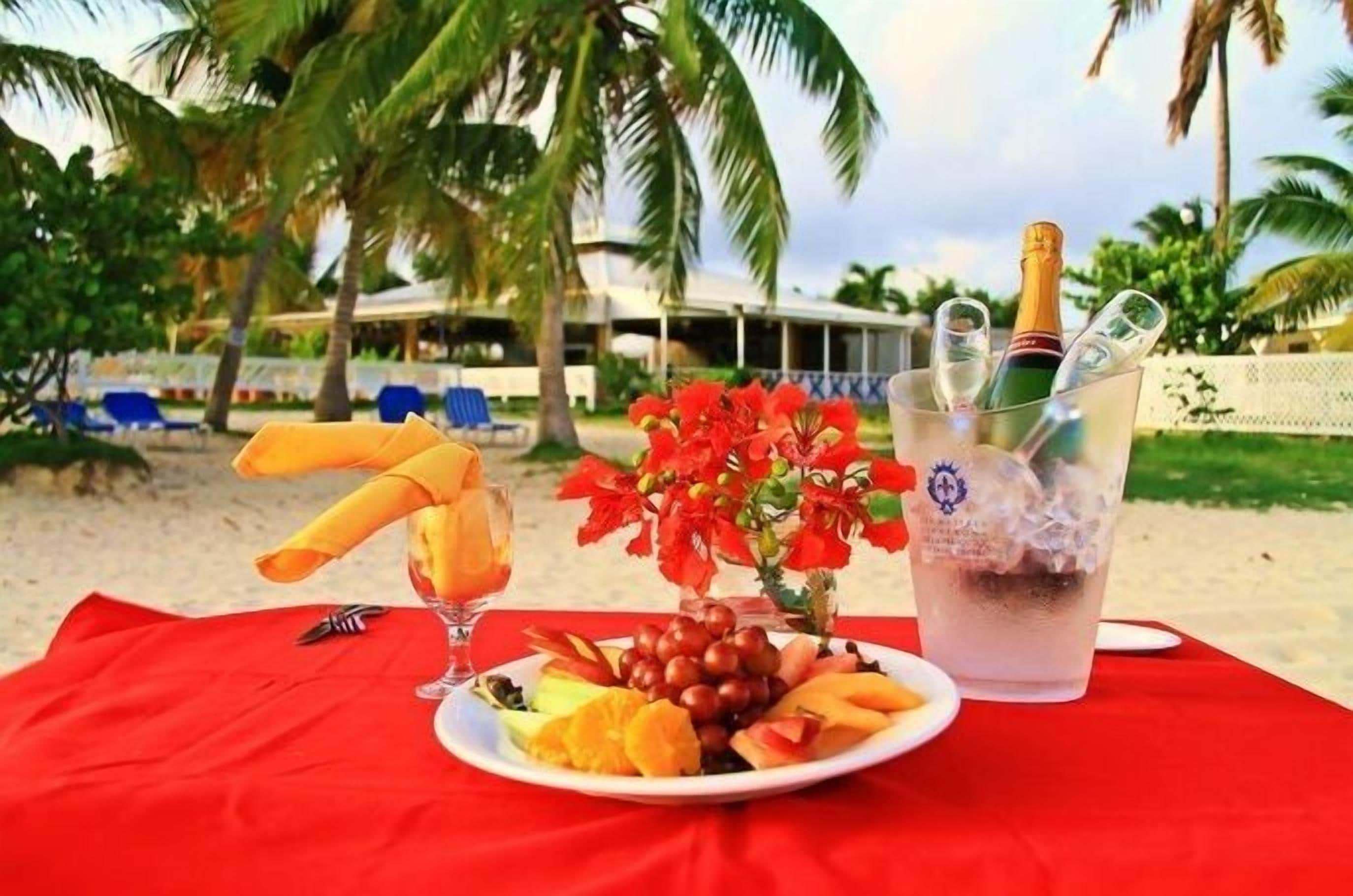 Une table installée sur la plage, avec une assiette de fruits, un verre de jus, une bouteille de champagne et un bouquet de fleurs.