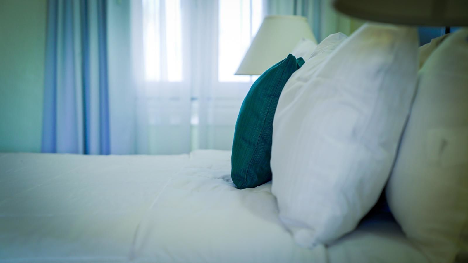 A neatly made bed with white and teal pillows, a lamp on the side, and a window with curtains in the background.