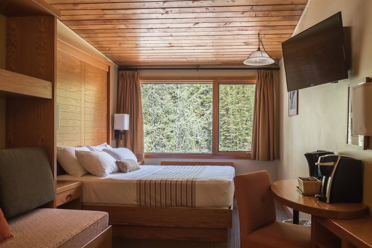 Una habitación de cabaña de madera con una cama, un escritorio, una silla y un ventanal con cortinas.