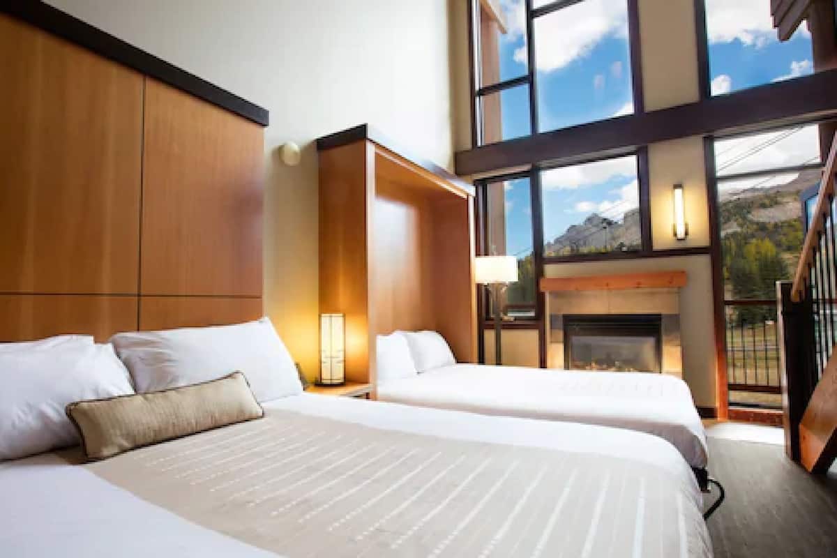 Habitación de hotel con dos camas, chimenea y ventanas amplias que dan a una vista de montaña.