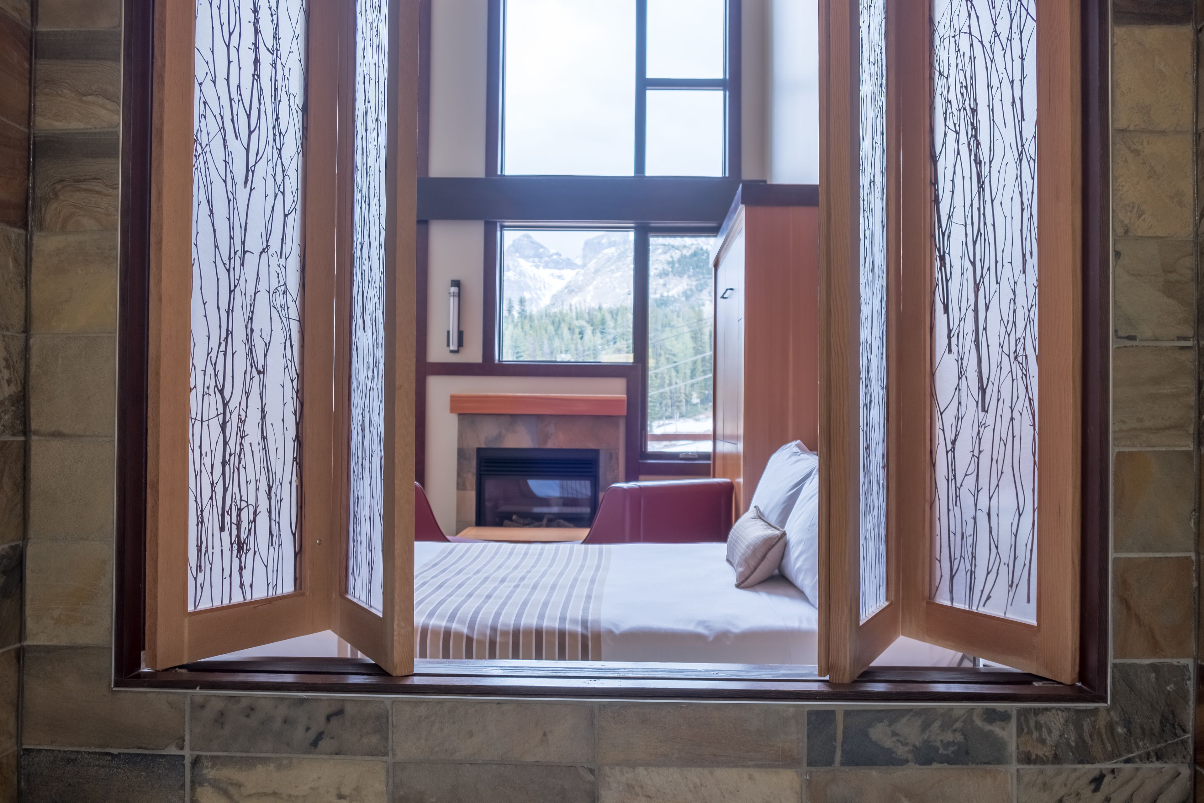 Un dormitorio con chimenea de piedra, una cama con almohadas y vistas a montañas a través de una ventana enmarcada por paneles de madera.