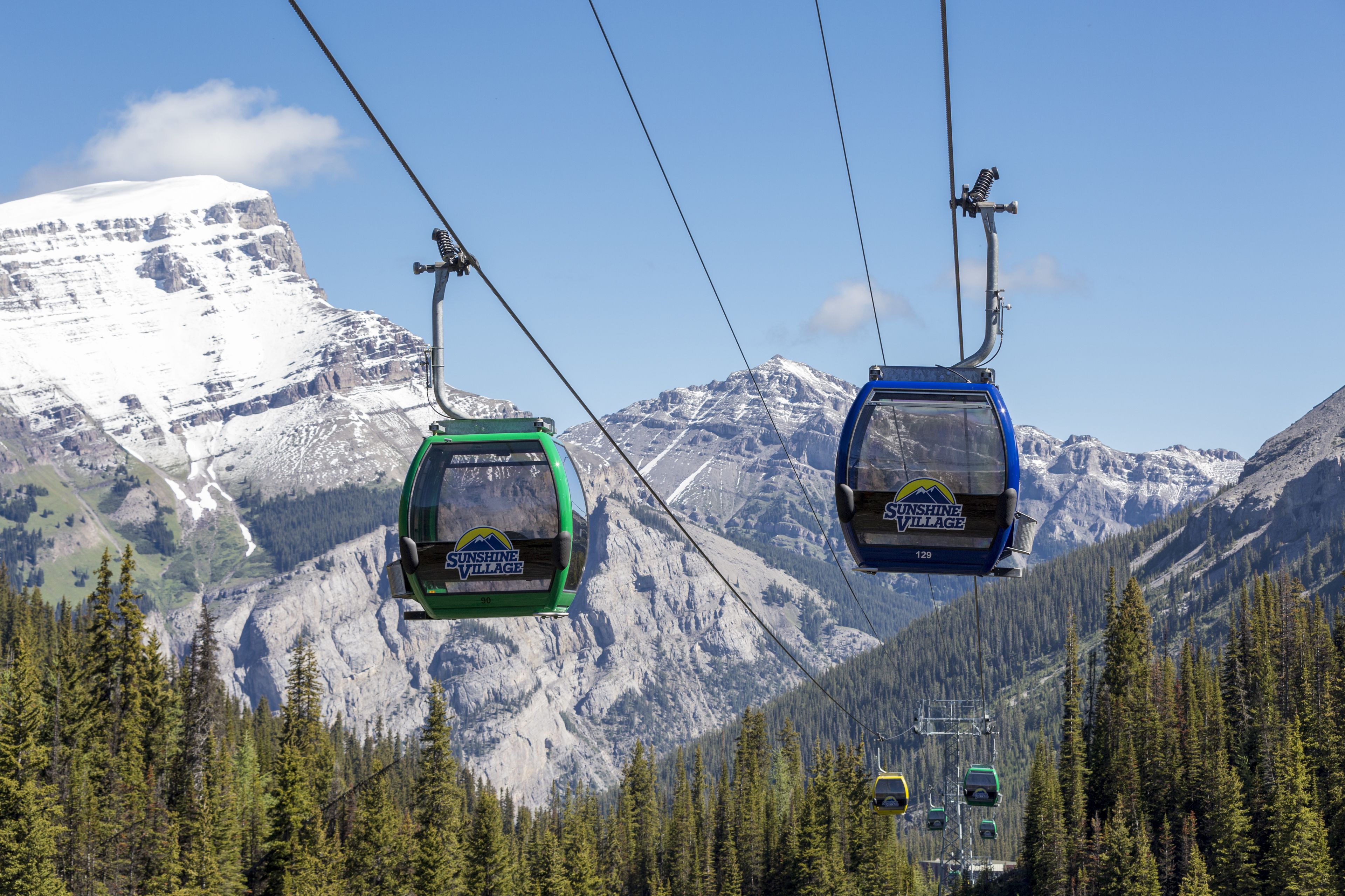 Two cable cars suspended above a mountainous landscape with snow-capped peaks and dense forests.