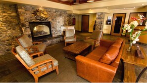 A cozy lobby with a stone wall, a fireplace, leather sofa, and wooden chairs.