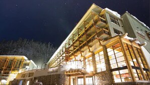 A multi-story building with a wooden exterior and large windows, surrounded by snow. - Sunshine Mountain Lodge (Sunshine)