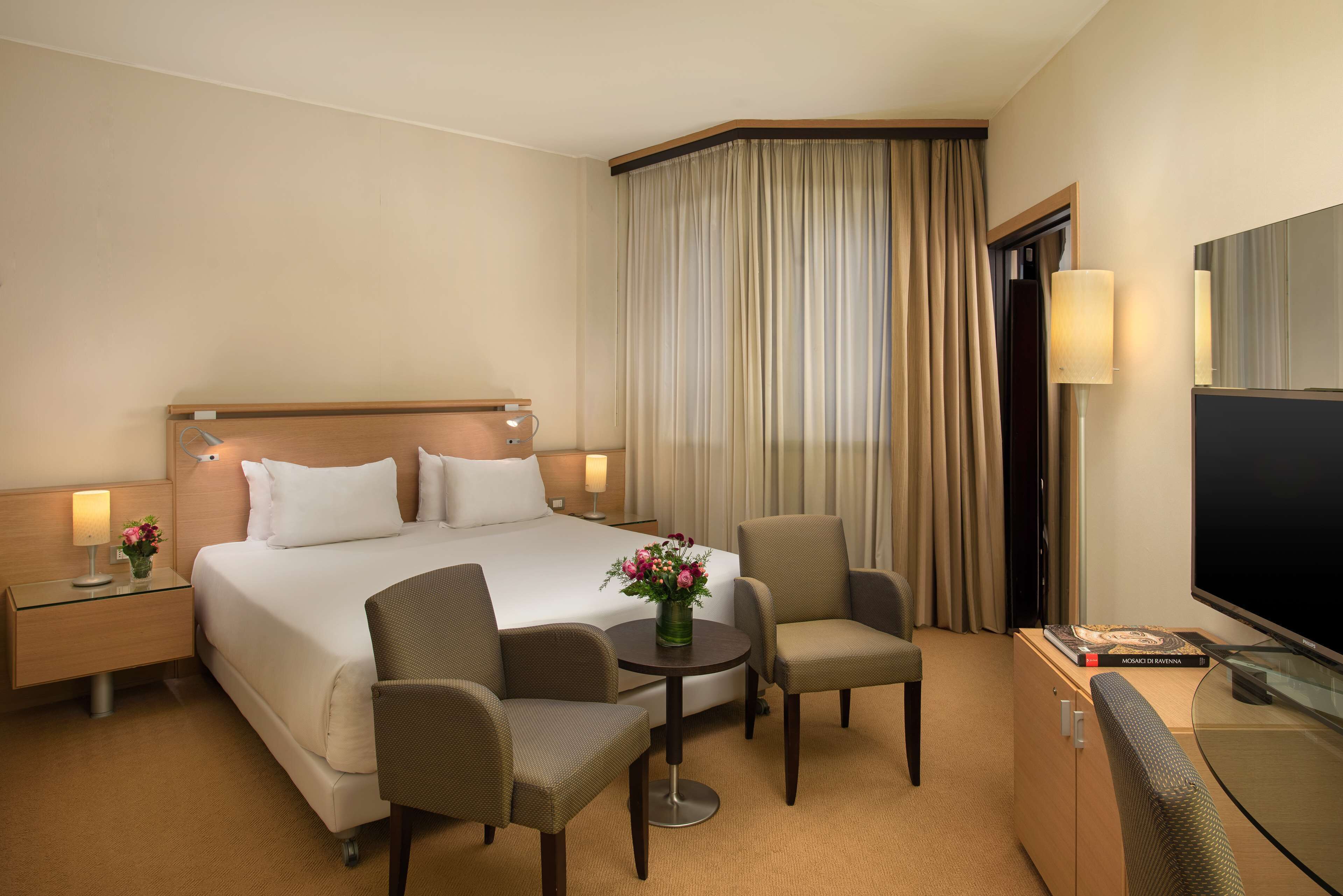 A hotel room with a large bed, two armchairs, a small table with a vase of flowers, a TV, and a minibar.
