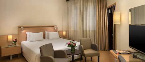 A hotel room with a large bed, two armchairs, a small table with a vase of flowers, a TV, and a minibar.