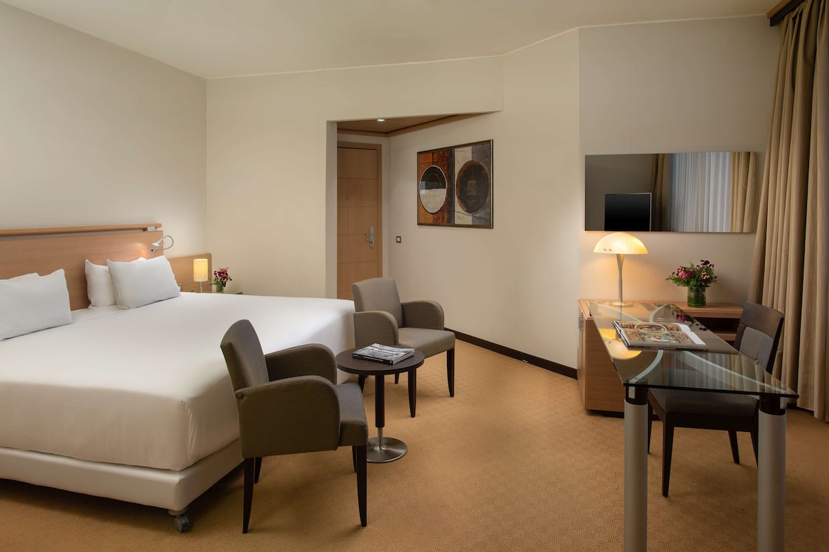 A hotel room with a large bed, two armchairs, a small table, a desk, and a TV.