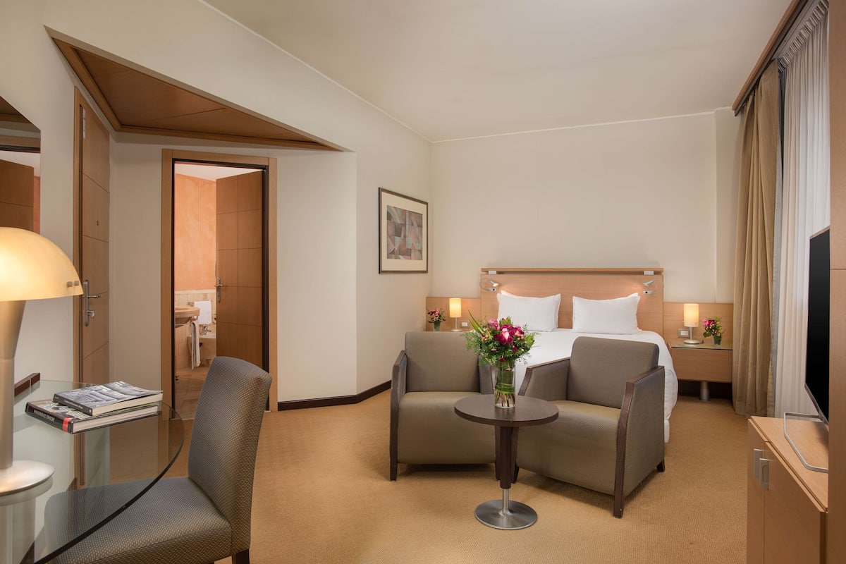 A hotel room with a bed, two armchairs, a small table with a vase of flowers, a bedside table with a lamp, and a television.