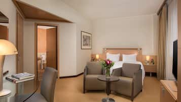 Habitación de hotel con una cama, dos sillones, una mesita con un jarrón de flores, una mesita de noche con una lámpara y un televisor.