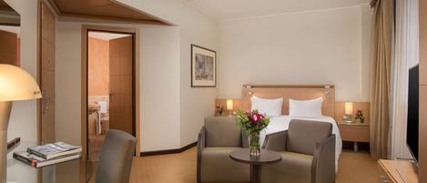 Habitación de hotel con una cama, dos sillones, una mesita con un jarrón de flores, una mesita de noche con una lámpara y un televisor.