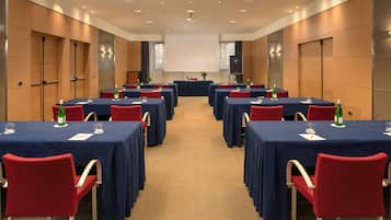 Ein Konferenzraum mit mehreren Tischen, roten Stühlen und einer großen Leinwand im Vordergrund.