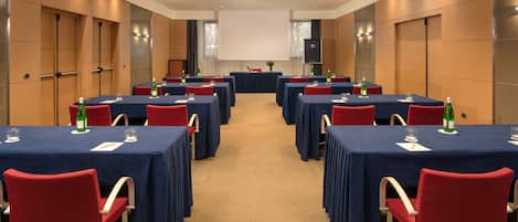 A conference room with multiple tables, red chairs, and a large screen at the front.