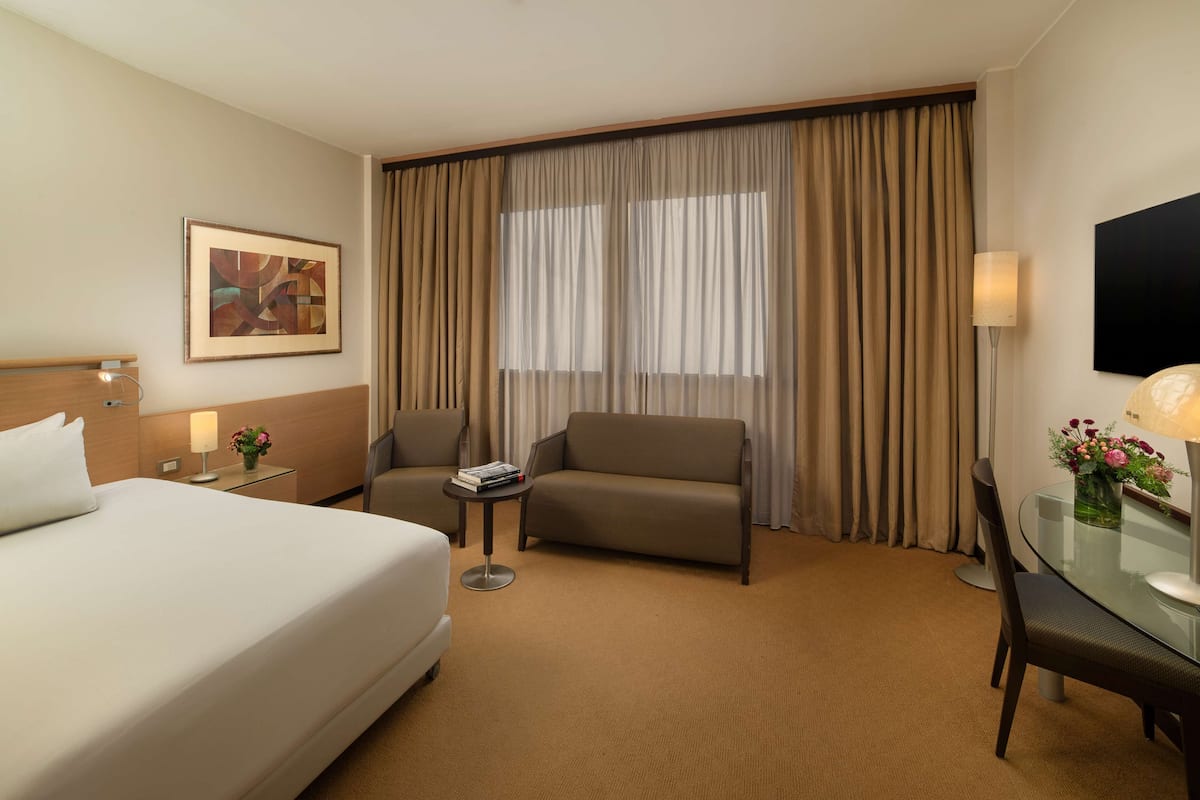 A hotel room with a bed, a sofa, a small table with a lamp, a chair, a TV, and a window with curtains.