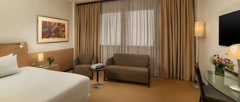 Une chambre d’hôtel avec un lit, un canapé, une petite table avec une lampe, une chaise, une télévision et une fenêtre avec des rideaux.