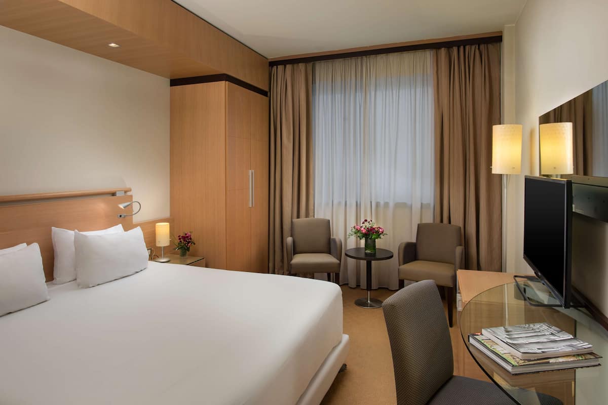 A hotel room with a large bed, two armchairs, a small table with a lamp, a TV, and a window with curtains.
