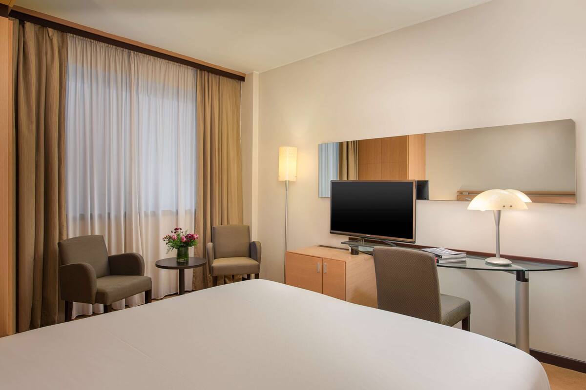 A hotel room with a large bed, a desk with a lamp, a television, and two armchairs.