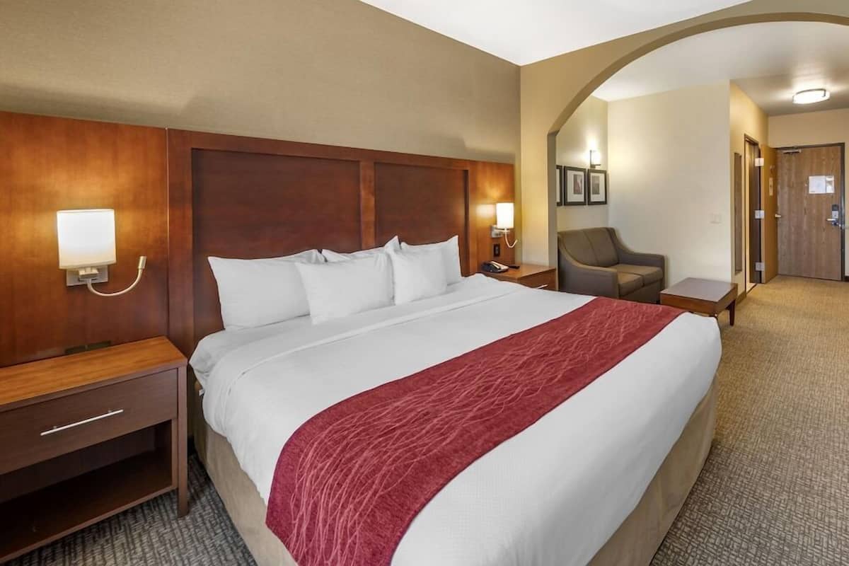 A hotel room with a large bed, a wooden headboard, a red throw blanket, a nightstand with a lamp, and a chair.