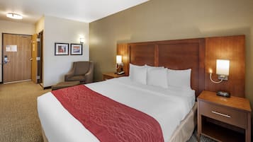 Quarto de hotel com uma cama grande, uma cadeira, uma mesa de cabeceira com abajur e uma porta.