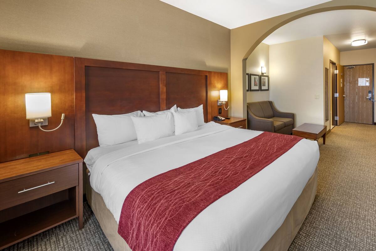 A hotel room with a large bed, a wooden headboard, a red throw blanket, a nightstand with a lamp, and a chair.
