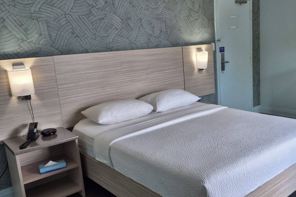 A hotel room with a bed, bedside lamps, a nightstand, a desk, and a trash bin.